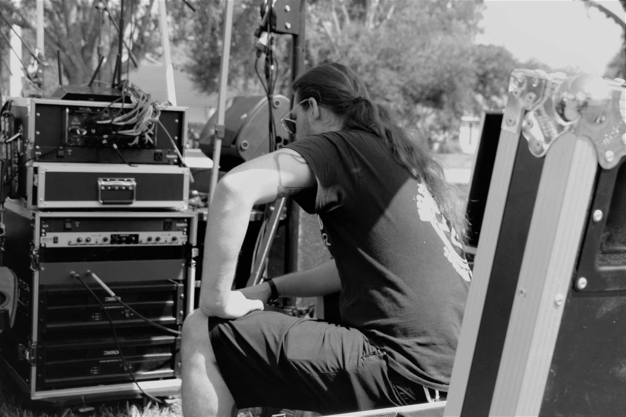 A black and white photo of a technician working at a rack of amplifiers and audio gear at an outdoor event.
